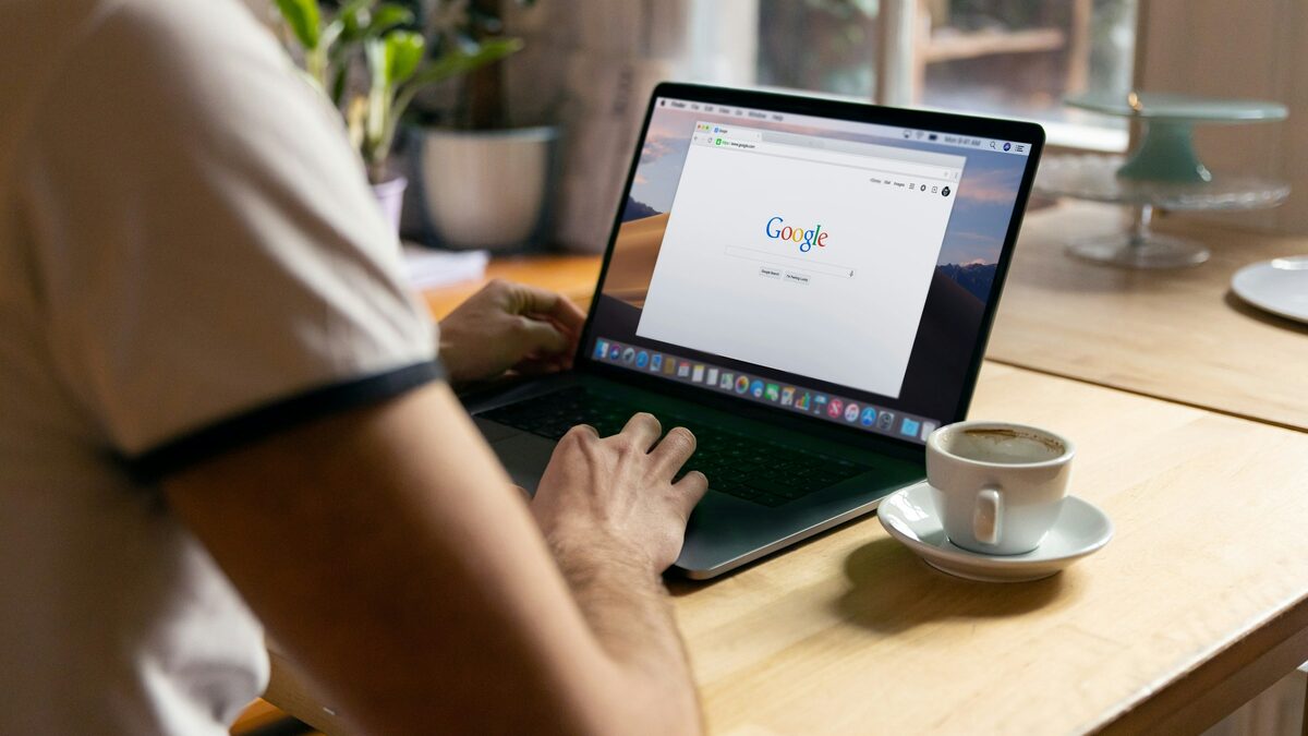 Person sitzt mit Kaffee am Holztisch vor einem Laptop mit der geöffneten Google-Suche
