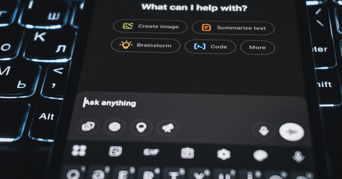 Smartphone vor einer Tastatur, auf dem Bildschirm die Eingabemaske eines KI-Assistenten mit der Frage „What can I help with?“
