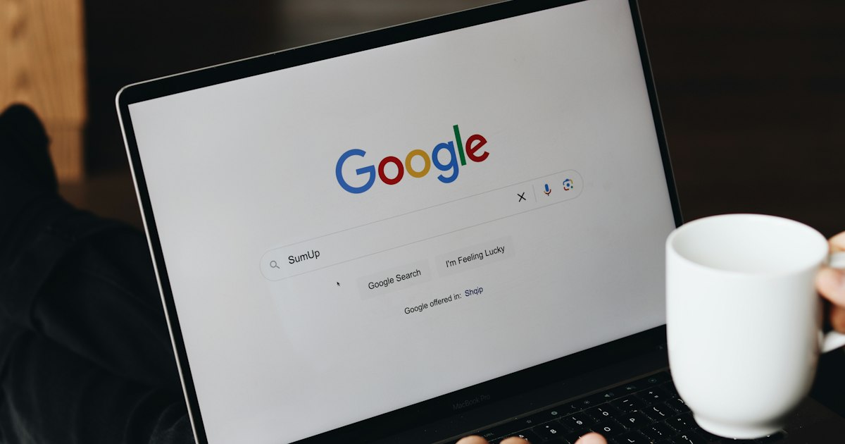 Person tippt an einem Laptop, auf dem Bildschirm die Google-Startseite, daneben eine Kaffeetasse