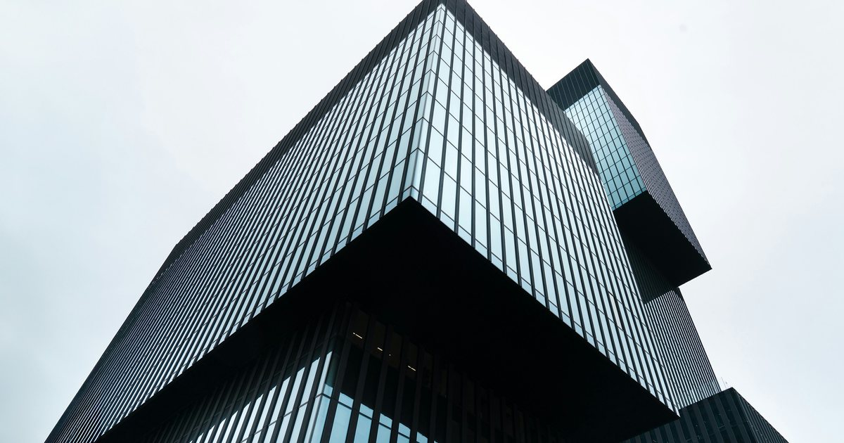 Modernes Bürogebäude mit dunkler Glasfassade und auskragendem Obergeschoss, von unten gegen hellen Himmel fotografiert